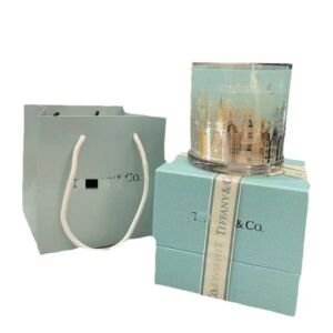 An isolated studio product shot featuring a glass scented candle decorated with a colorful city skyline illustration, resting atop a light teal textured gift box wrapped with a branded ribbon alongside a matching paper gift bag on a plain white background.