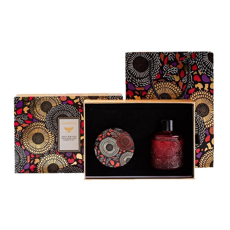 An isolated product shot of a luxury fragrance gift set featuring a deep red embossed glass reed diffuser bottle, a floral tin scented candle, a matching presentation box, and a printed gift bag with a vibrant sunflower motif against a white background.