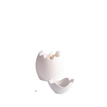 An isolated studio product shot of a two-piece matte white ceramic candle holder set shaped like a cracked eggshell, featuring a lit tealight flame inside the main vessel and a smaller matching wavy-edged dish in front against a clean white background.