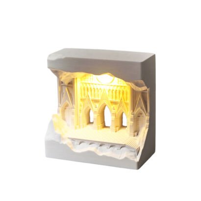 A white rectangular resin sculpture carved to reveal an illuminated interior scene of Gothic arches and cathedral facades emitting a warm golden LED glow against a plain white background.