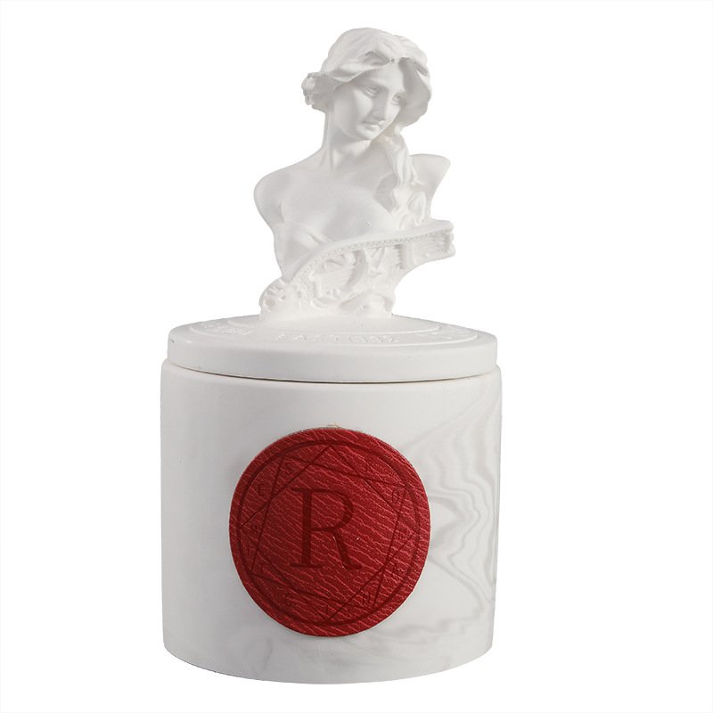 A matte white ceramic scented candle jar featuring a sculpted Classical Greek goddess bust handle on the lid and a round red wax-seal style medallion stamped with the letter R on the front.