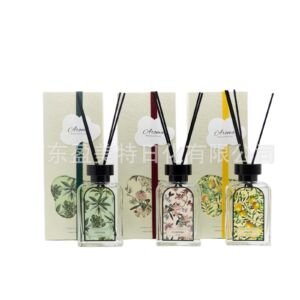 A set of three clear glass reed diffusers featuring leafy, floral, and citrus botanical label prints, placed in front of matching ribbon-accented packaging boxes.