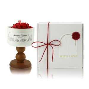 A red rose-shaped carved soy candle housed in a white ceramic goblet with a turned wooden pedestal stand, set next to a white tied gift box with a red wax seal.