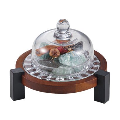 A clear glass dome cover over raw green crystal stones, cinnamon sticks, and dried botanicals resting on a glass dish set within a round wooden tripod stand.