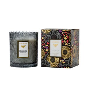 A luxury scented soy candle in an embossed dark smoky glass vessel next to a vibrant floral and bee patterned gift box.
