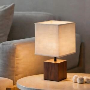 A modern minimalist table lamp with a solid cubic dark wood base and a square off-white fabric shade emitting a warm cozy glow, placed on a marble side table beside a grey sofa.
