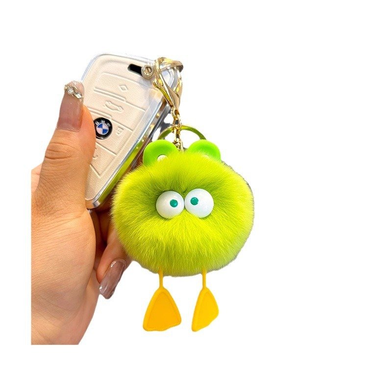 A vibrant lime green plush pom pom keychain with big googly eyes, green frog-like ears, and dangling yellow webbed duck feet, attached to a car key fob.