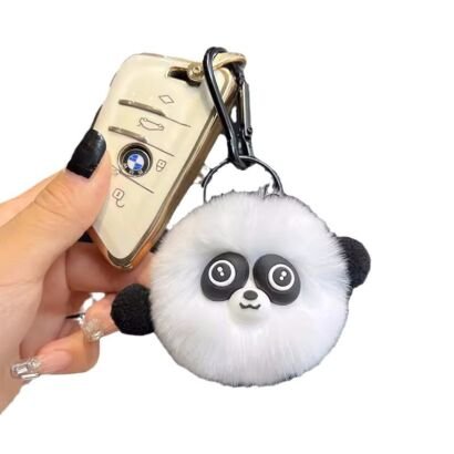 A hand holding a white fluffy pom-pom panda keychain next to a white BMW car key fob, featuring black eye patches with wide round eyes, black plush ears, a cute small snout, and a black clip clasp.