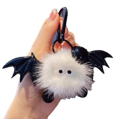 A hand holding a white fluffy pom-pom bat keychain featuring glossy black faux leather wings, small black bead eyes, tiny black feet, and a black carabiner clip clasp against a clean background.