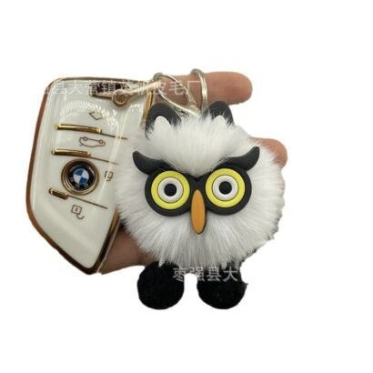 A hand holding a white fluffy pom-pom owl keychain next to a white BMW car key fob, featuring stylized yellow owl eyes with black trim, an orange beak, black ear tufts, and tiny black feet.
