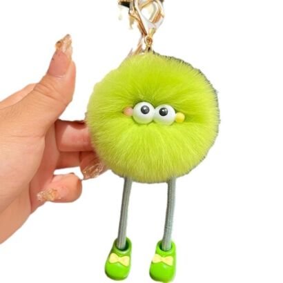 A vibrant lime green fluffy pom-pom monster keychain charm held in a hand, featuring big round googly eyes, rosy cheeks, dangling cord legs, cute green shoes with yellow bows, and a gold metal clip clasp.