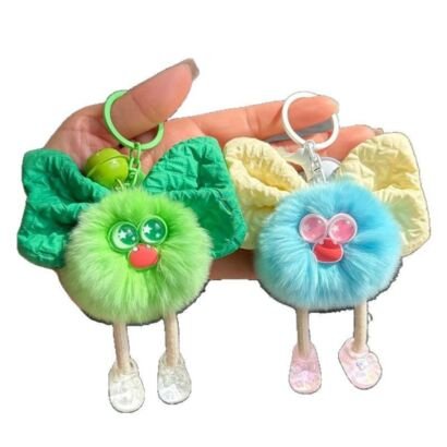 A hand holding two fluffy plush monster keychains—one vibrant green with a matching green fabric bow, and one pastel blue with a light yellow fabric bow—both featuring large cartoon eyes, red lips, dangling legs, and small clear shoes.