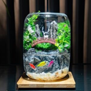 A curved glass vessel featuring a dual-level display: a aquatic bottom level with colorful fish swimming among light pebbles and a rock backdrop, and a lush land top level with a miniature arched bridge, a white deer figurine, green moss, plants, and white flowers, resting on a wooden base.