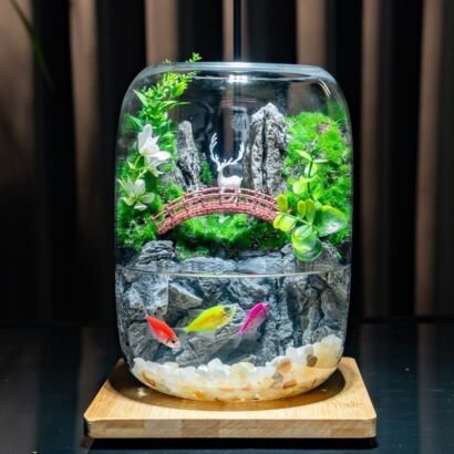 A curved glass vessel featuring a dual-level display: a aquatic bottom level with colorful fish swimming among light pebbles and a rock backdrop, and a lush land top level with a miniature arched bridge, a white deer figurine, green moss, plants, and white flowers, resting on a wooden base.