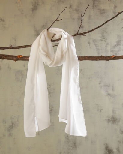 A lightweight, semi-sheer off-white silk chiffon scarf draped gracefully over a natural wooden tree branch against a textured neutral wall backdrop.