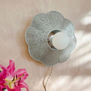 A modern flower-shaped wall sconce featuring a scalloped metallic woven thread shade and a central frosted ribbed round light bulb mounted on a natural light wood wall.