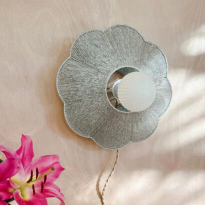 A modern flower-shaped wall sconce featuring a scalloped metallic woven thread shade and a central frosted ribbed round light bulb mounted on a natural light wood wall.