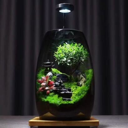 A tinted glass terrarium container resting on a wooden base, featuring a lush green miniature tree, mossy hills, a small pagoda, red foliage accent, stone steps, and an overhead LED grow light against a dark curtain backdrop.