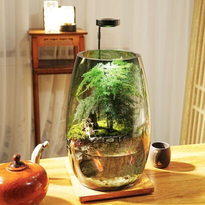A tall, oval glass jar desktop ecosystem illuminated by an overhead LED light, featuring a lush green asparagus fern and moss landscape on top and a small aquatic section with swimming goldfish below, set on a wooden table.