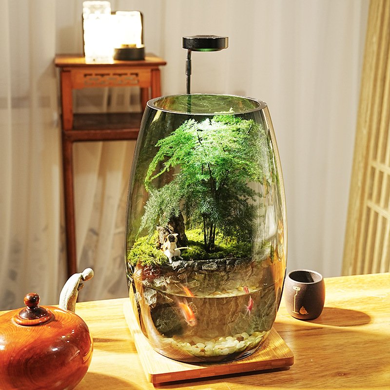 A tall, oval glass jar desktop ecosystem illuminated by an overhead LED light, featuring a lush green asparagus fern and moss landscape on top and a small aquatic section with swimming goldfish below, set on a wooden table.